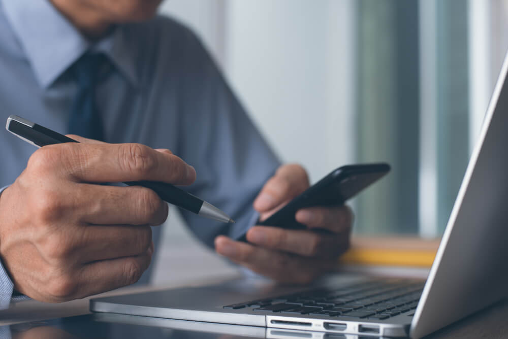 Businessman, lawyer or accountant holding a pen and using mobile smart phone working on laptop computer on office desktop, close up, browsing internet on electronics device