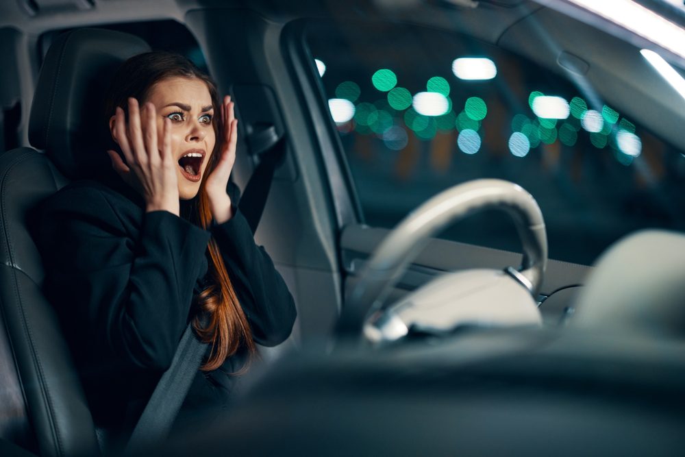 A women sitting in the drivers seat of a car, holds he hands to her face in shock.