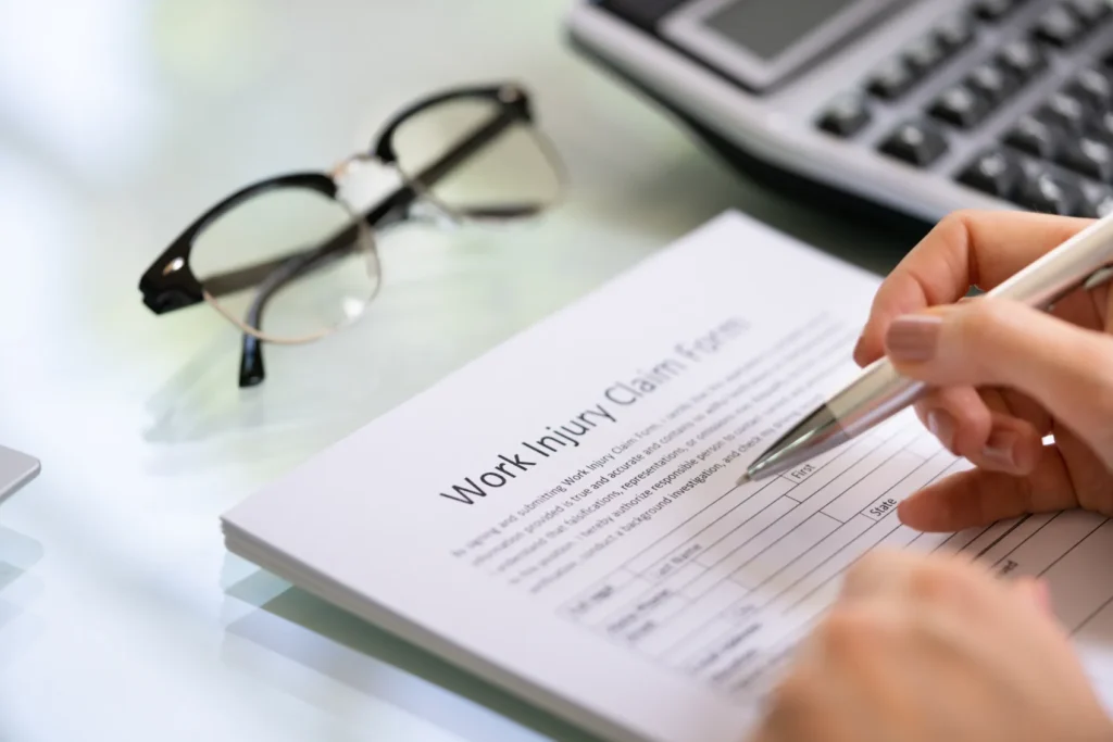 A person filling out a work injury form with a pair of eyeglasses rested on the table.